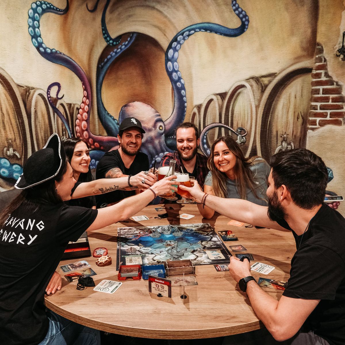 Friends raising their beers at a board game table under the octopus mural